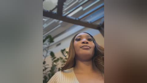 Media: Video of a confident, light-skinned Black woman with long, wavy brown hair, wearing a cream-colored, ribbed sweater, standing in an indoor setting with a green plant wall and industrial ceiling.