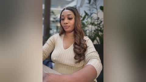 Media: Video of a Black woman with long wavy brown hair, wearing a cream-colored knit sweater, sitting outdoors with blurred greenery in the background.