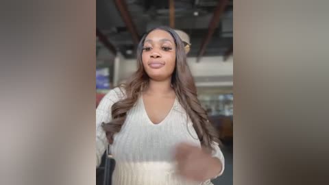 Media: Video of a confident Black woman with long, wavy hair, wearing a beige sweater and beige hat, standing in a modern, industrial setting with exposed pipes.