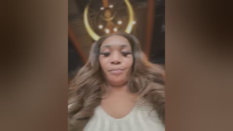 Media: A video of a young Black woman with long, wavy brown hair and a golden horn-like headdress. She wears a white top, indoors with a blurred background.