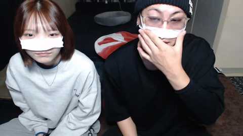 Media: Video of two young Asian women with white tape over their mouths, wearing matching black beanie hats and sweatshirts, sitting on a carpeted floor in a dimly lit room.