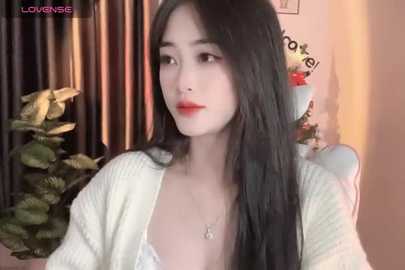 Media: Video of an East Asian woman with long black hair, fair skin, and red lipstick, wearing a cream-colored cardigan, seated indoors with a plant and wall decor in the background.