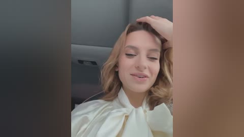 Media: Video of a young woman with light skin and wavy blonde hair, wearing a white blouse, sitting in a car. She has a relaxed expression, with one hand on her head, and the background shows car interior details.