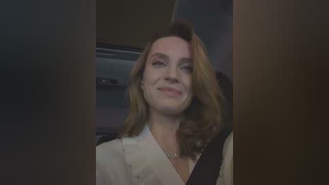 Media: Video of a Caucasian woman with shoulder-length blonde hair, wearing a white blouse and a seatbelt, smiling slightly. Background shows a dimly lit interior, possibly a car or an airplane.