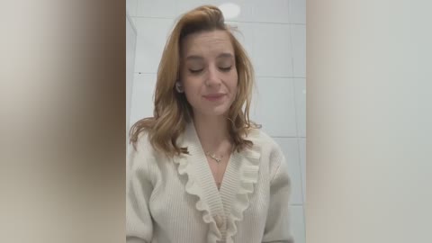 Media: Video of a young, fair-skinned woman with shoulder-length, wavy, light brown hair, wearing a cream-colored ruffled blouse, standing in a tiled bathroom. She has her eyes closed and a slight smile.