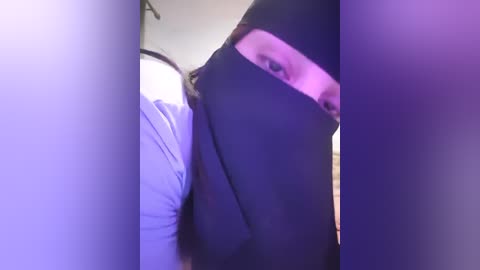 Media: A video of a person with light skin and brown hair partially obscured by a black mask, wearing a white shirt, taken indoors under purple lighting.