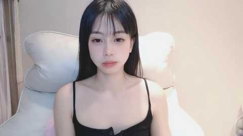 Media: A video of a young Asian woman with long black hair, wearing a black spaghetti strap top, sitting on a white cushioned chair against a plain light gray wall.