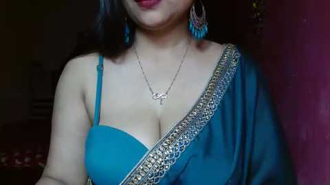 Media: A video of a light-skinned woman with shoulder-length dark hair, wearing a blue saree with gold embroidery, a silver necklace, and turquoise earrings, partially visible. The background is dimly lit with a dark wall.