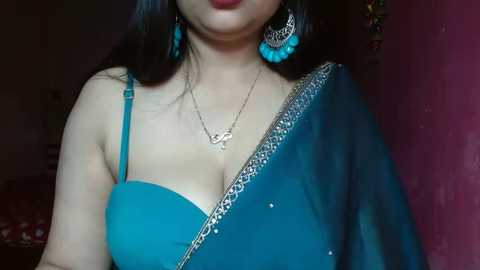 Media: Video of a fair-skinned woman with long black hair, wearing a turquoise blue sari with a plunging neckline, silver jewelry, and a maroon wall background.