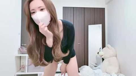 Media: Video of an Asian woman with long brown hair, wearing a black long-sleeve top and white mask, bending over in a bedroom with a wooden wardrobe, teddy bear, and a cluttered nightstand.