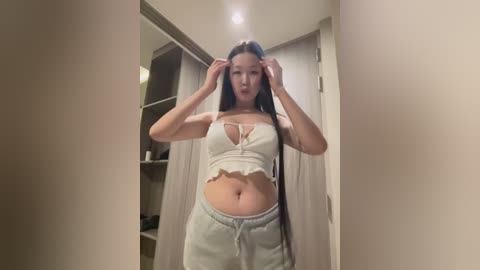 Media: Video of an Asian woman with long black hair, wearing a white cropped top and light gray sweatpants, adjusting her hair in a hallway with beige walls and closed doors.