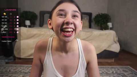 Media: Video of a smiling Asian woman with dark hair, wearing a white tank top, in a modern bedroom with beige walls, a bed, and potted plants.