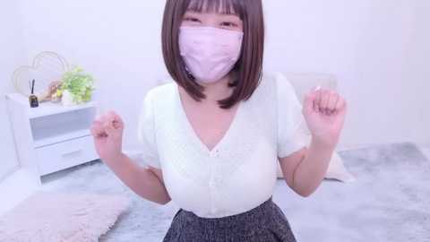 Media: A young Asian woman with a bob haircut, wearing a pink mask, a white blouse, and a pleated skirt, poses playfully with clenched fists in a minimalist, light-colored room.