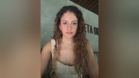 Media: A video of a young woman with curly, light brown hair, fair skin, and brown eyes, wearing a sleeveless top, standing indoors with a concrete wall and wooden ceiling in the background.