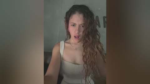 Media: A video of a young woman with long, curly hair, wearing a beige tank top, seated indoors, looking surprised, with a blurred background.