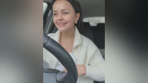 Media: Video of a smiling, light-skinned woman with dark hair, wearing a white jacket, driving a car with a black steering wheel.