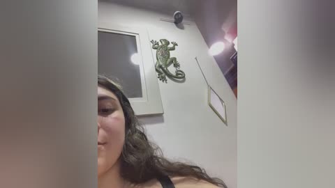 Media: Video of a young woman with long, wavy brown hair, wearing a black tank top, captured from a low angle, showing a close-up of her face. Background features a white wall with a green frog statue and a window.