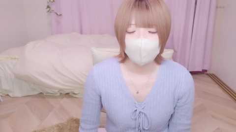 Media: A video shows a light-skinned woman with short blonde hair, wearing a light blue ribbed top and a white face mask, sitting on a wooden floor in a softly lit room with a bed and pastel-pink curtains.