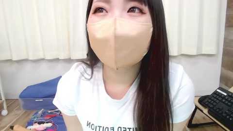 Media: Video of an East Asian woman with long black hair, wearing a white t-shirt and a beige surgical mask, standing indoors with a blue chair and a black keyboard in the background.