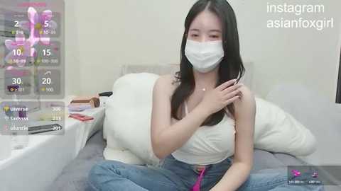 Media: A young Asian woman with long black hair, wearing a white mask and a white top, sits cross-legged on a bed in a bedroom, adjusting her hair. A digital overlay shows Instagram stats.