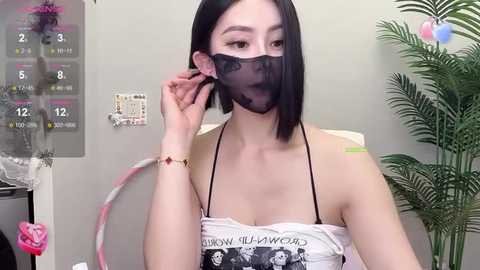 Media: Video of a young East Asian woman with straight black hair, wearing a black face mask and a white top with a black floral print. Background includes a calendar, plant, and wall decor.