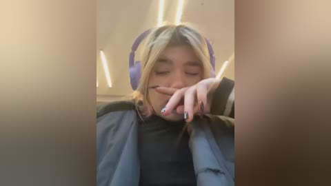 Media: A video of a young woman with long blonde hair, wearing large purple headphones, resting her head on a table, eyes closed, and hand covering her mouth.