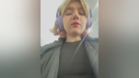 Media: Video of a young person with short, blonde hair wearing large, purple headphones and a black jacket, sitting in a dimly lit room, eyes closed.