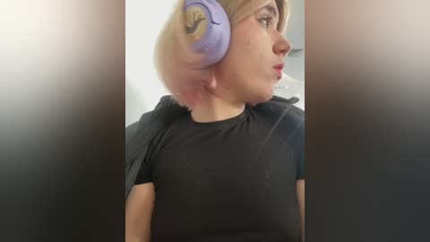 Media: A video of a fair-skinned woman with blonde hair in a ponytail, wearing large, pastel-colored headphones and a black T-shirt, seen from the side.