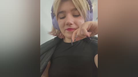 Media: Video of a young woman with fair skin, short blonde hair, wearing purple headphones and a black top, leaning against a blurred background, with a person's hand gently touching her chin.