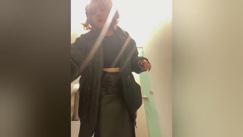 Media: A video of a person with a short, wavy blonde bob haircut, wearing a black coat and pants, walking through a dimly lit hallway with beige walls and a green door.