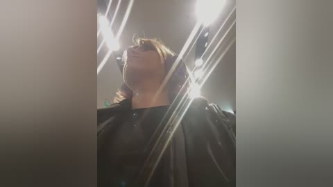 Media: Video of a person with light skin and long, straight brown hair, wearing glasses and a black plastic cape, standing in a dimly lit room with multiple bright spotlights creating a dramatic effect.