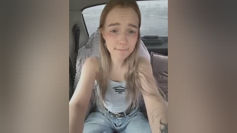 Media: Video of a young, fair-skinned woman with long, blonde hair, wearing a white tank top and light blue jeans, sitting in a car, looking slightly sad.