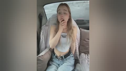 Media: Video of a young woman with long blonde hair, wearing a white crop top and jeans, sitting in a car with a distressed expression, fingers to her mouth.