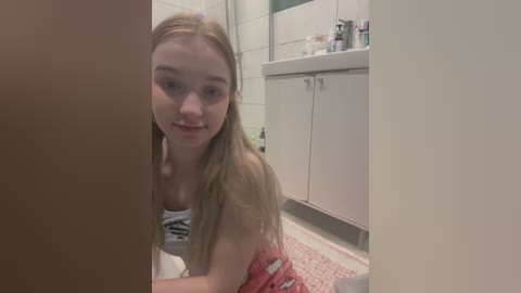 Media: Video of a young blonde woman with fair skin, wearing a white tank top, kneeling on a pink rug in a bathroom. White cabinets and toiletries are visible in the background.