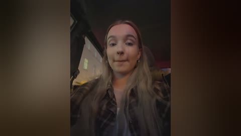 Media: Video of a young woman with long, blonde hair, wearing a dark jacket, sitting in a dimly lit car interior, looking straight at the camera.