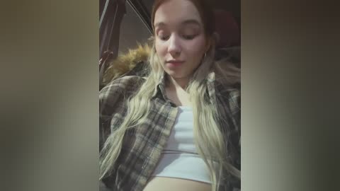 Media: Video of a young woman with long, straight blonde hair, wearing a white crop top and plaid shirt, sitting in a car, eyes closed, smiling softly.