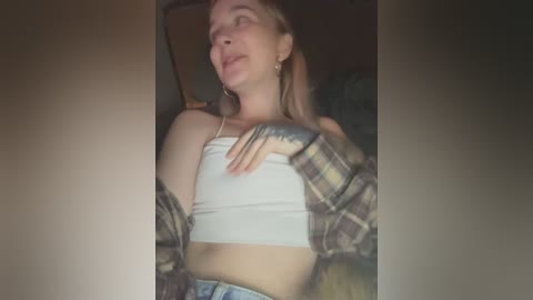 Media: Video of a young woman with fair skin, light brown hair, wearing a white crop top and plaid shirt, lying on a bed, eyes closed, arm on stomach.