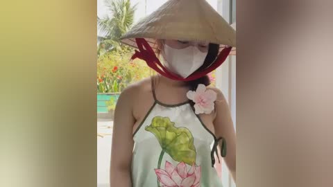 Media: Video of an Asian woman wearing a traditional conical hat, face mask, and sleeveless floral blouse. She stands indoors with a tropical garden visible through a window, featuring green leaves and red flowers.