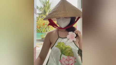 Media: Video of a woman in a Vietnamese conical hat, wearing a floral blouse, mask, and sunglasses, standing in a garden with tropical plants.