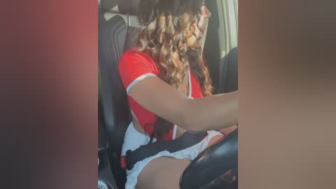 Media: Video of a young woman with wavy brown hair, wearing a red shirt and white shorts, seated in a car, driving.