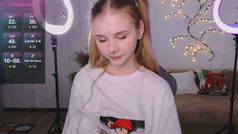 Media: Video of a young girl with light skin and blonde hair, wearing a white T-shirt with a graphic, sitting indoors. Background includes a bed, fairy lights, and a ring light.