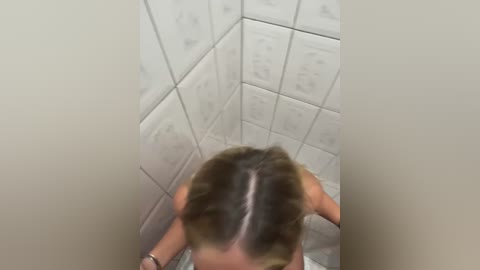 Media: A video of a woman with long, blonde hair, wet and slicked back, standing in a tiled shower. The walls are covered with white tiles, and the lighting is dim.