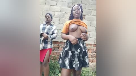 Media: Video of two Black women standing outdoors, one lifting her shirt to expose her breast, wearing a plaid shirt and skirt, the other wearing a checkered shirt and red skirt. Background includes a brick wall and greenery.