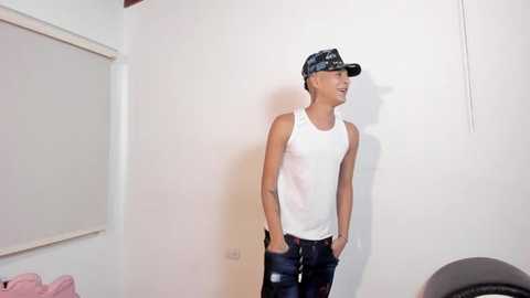 Media: Video of a young man in a white tank top, dark jeans, and a black baseball cap, standing against a plain white wall with a pink couch and blinds in the background.