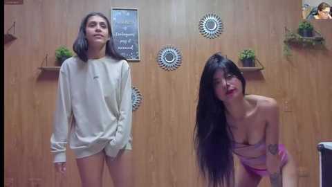 Media: Video of two women with long black hair in a wooden room; one wears a loose sweater, the other a strapless top.