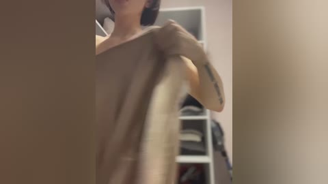 Media: Video of a woman with short dark hair, holding a brown towel, revealing a tattoo on her right forearm, in a dimly lit room with shelves in the background.