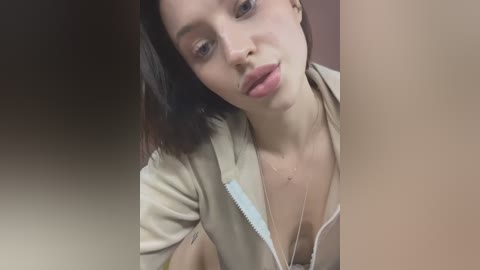 Media: Video of a young woman with fair skin and straight brown hair, wearing a beige jacket and a silver necklace. She has a neutral expression. The background is blurred.