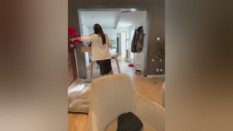 Video of a woman with long hair, wearing a white shirt, standing in a modern, minimalistic apartment with beige walls, wooden floors, and a partially open door revealing a hallway.