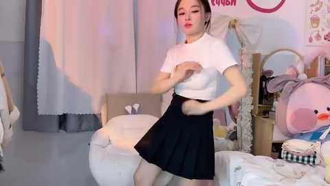 Media: Video of an Asian woman in a white crop top, black skirt, and pink lipstick dancing in a cozy, pastel-themed bedroom with stuffed animals and a white chair.