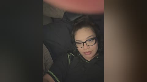 Media: A video of a woman with medium skin tone, short black hair, wearing glasses, and a black top, lying on a dark couch, partially obscured by a blurred object.